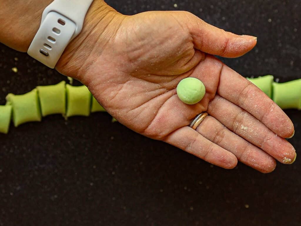 A hand holds a small, green dough ball, sprinkled with flour. In the background, a row of similarly colored dough pieces is lined up on a black surface. The person wears a white wristband and a ring.